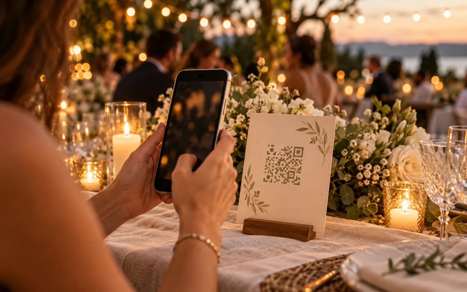 A wedding guest holds a phone toward a cream invitation card on a candlelit reception table, with flowers and guests softly blurred behind it.